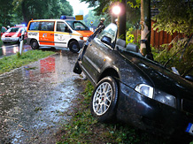 Am Rand der regennassen Fahrbahn steht ein schwarzer Audi mit abgetrenntem Dach halb auf dem Radweg, halb in angenzenden Wald leicht um einen Baum herumgebogen, im Hintergrund ein Notarzt-Einsatzfahrzeug und ein Feuerwehr-Einsatzleitwagen