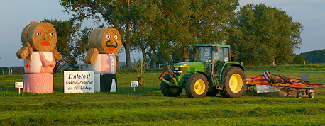 Ein Traktor mit einem angehngten Schwader passiert die zwei Erntefiguren auf einer Wiese